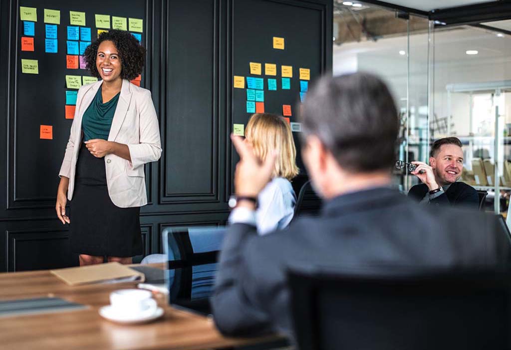 Woman at the front of a conference room discussing a project with co-workers.